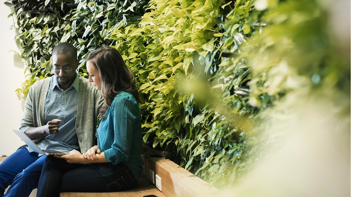 Businessman and woman sitting in front of green plant wall, using laptop