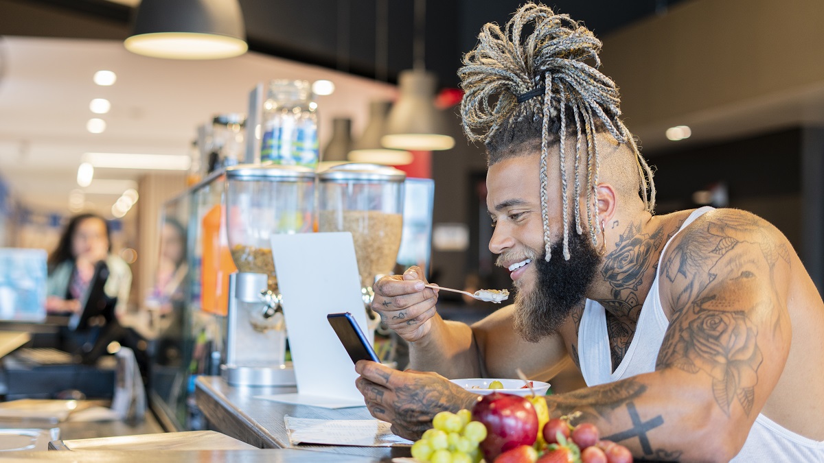 Latino man with an approximate age of 30 years is sitting at the gym food bar with a plate of salads in front of him a red apple in one of his hands and in the other his mobile phone which he checks with great concentration